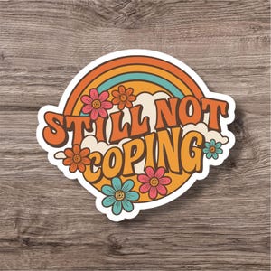 Może przedstawiać: Naklejka w stylu retro z napisem "STILL NOT COPING" w kolorze pomarańczowym. Projekt zawiera tęczę, chmury i kwiaty w kolorze różowym, pomarańczowym i turkusowym. Naklejka ma białą obwódkę i jest umieszczona na tle słojów drewna.