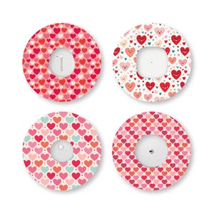 May include: Four circular adhesive patches for medical sensors, each featuring a distinct heart-themed pattern. The designs include various shades of red, pink, and teal hearts, with some hearts displaying simple smiling faces on a white background.