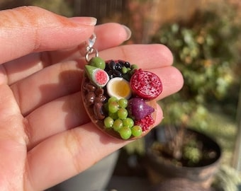 May include: A miniature food charm necklace featuring a wooden platter with a variety of fruits, including grapes, figs, and pomegranate. The charm is made of polymer clay and has a silver chain.
