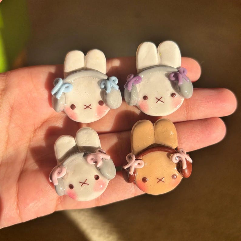 May include: Four handmade polymer clay bunny charms with different colored bows. The bunnies are white, gray, and brown with black eyes and a black nose.