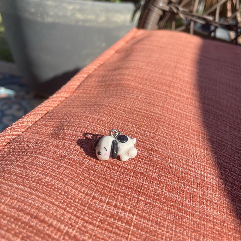 May include: A small, white and black ceramic dog charm with a silver loop. The dog is lying down with its eyes closed.