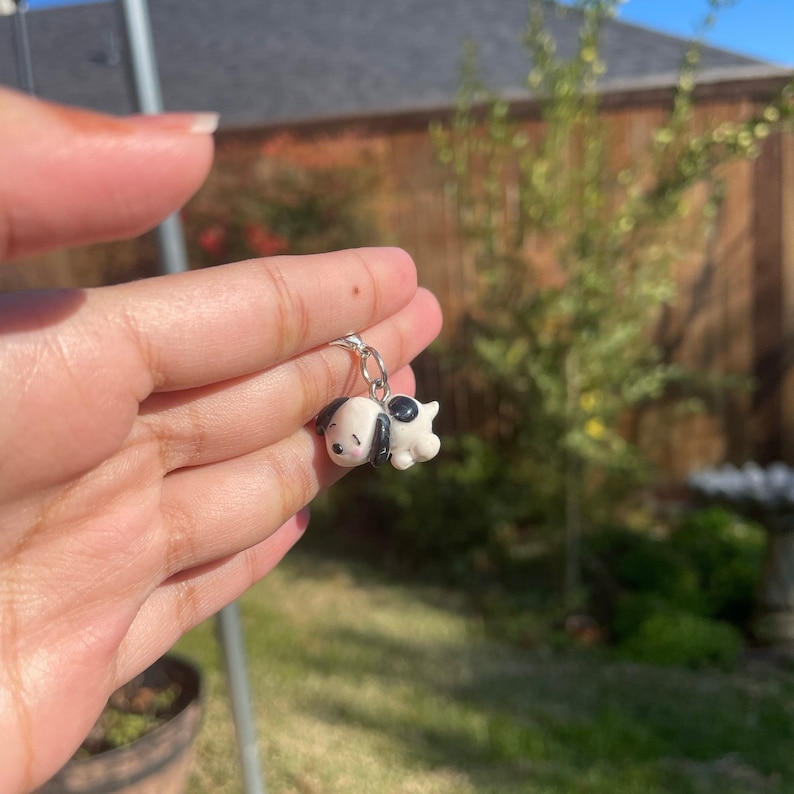 May include: A small white and black clay charm of a sleeping dog with a silver chain attached.