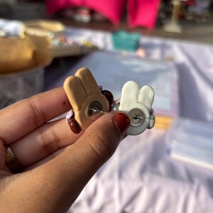 May include: A pair of brown and white bunny-shaped earrings with pink and purple accents. The earrings are made of polymer clay and have a simple, cute design.