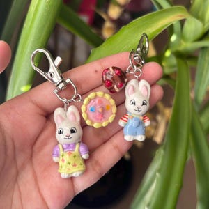 May include: A collection of handmade bunny-themed keychains. One bunny wears a yellow dress, another wears blue overalls. Additional charms include a miniature cake and a chocolate-covered treat. Each keychain has a silver clasp.