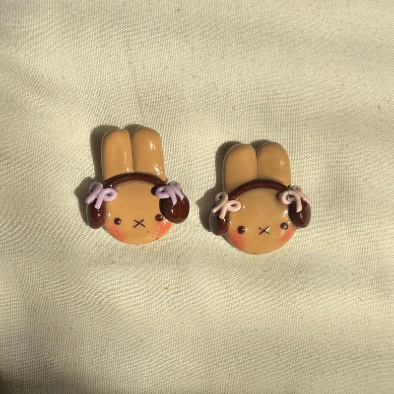 May include: Two brown bunny-shaped polymer clay earrings with brown ears and purple bows. The bunnies have pink cheeks and brown eyes.