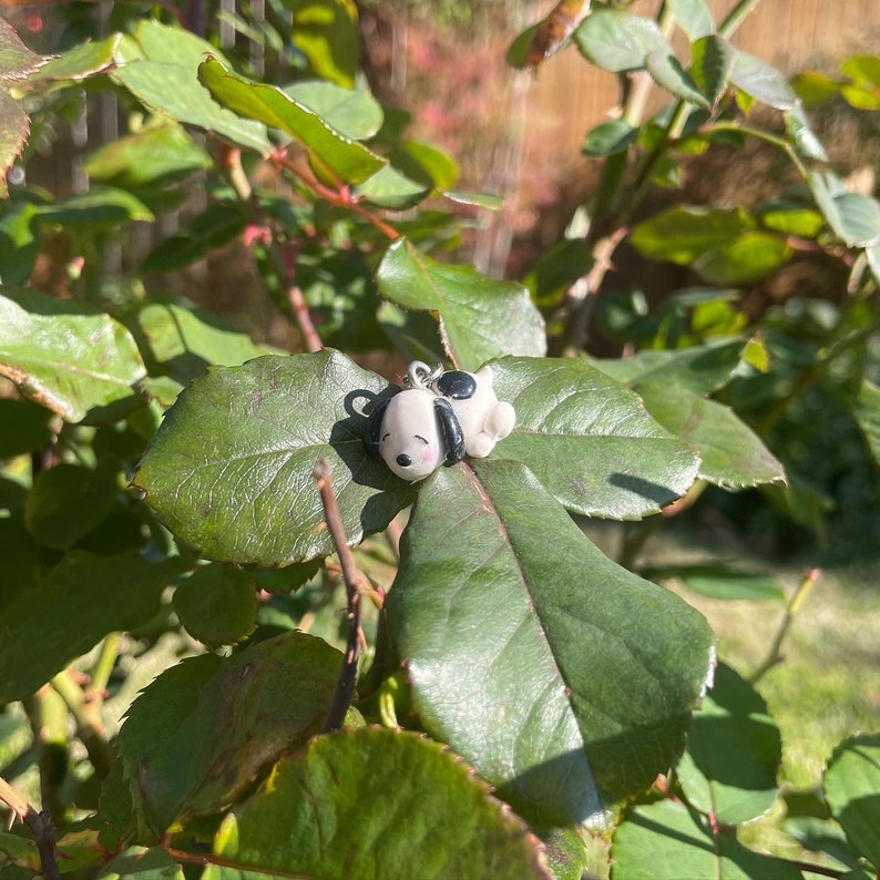 May include: A small white and black ceramic dog charm, lying on a green leaf. The dog has black eyes and a black nose, and is curled up in a sleeping position.