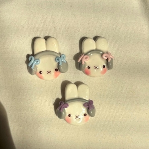 May include: Three white bunny figurines with gray headphones and colorful bows. The bunnies have pink cheeks and black eyes.