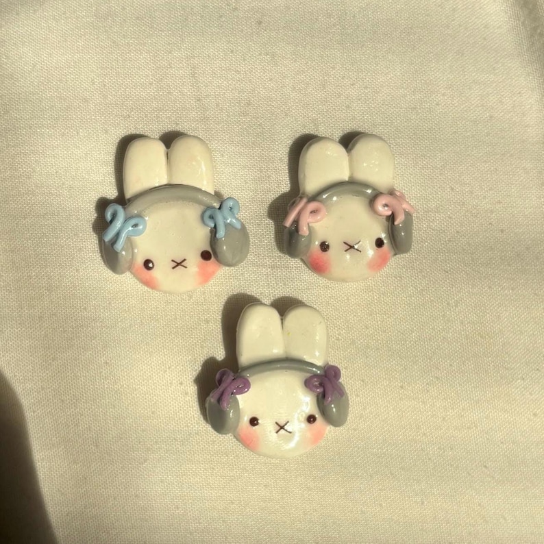 May include: Three white bunny figurines with gray headphones and colorful bows. The bunnies have pink cheeks and black eyes.