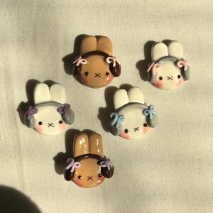 May include: Five handmade polymer clay bunny charms with pastel colored bows and gray or brown ears. The bunnies are wearing headphones.