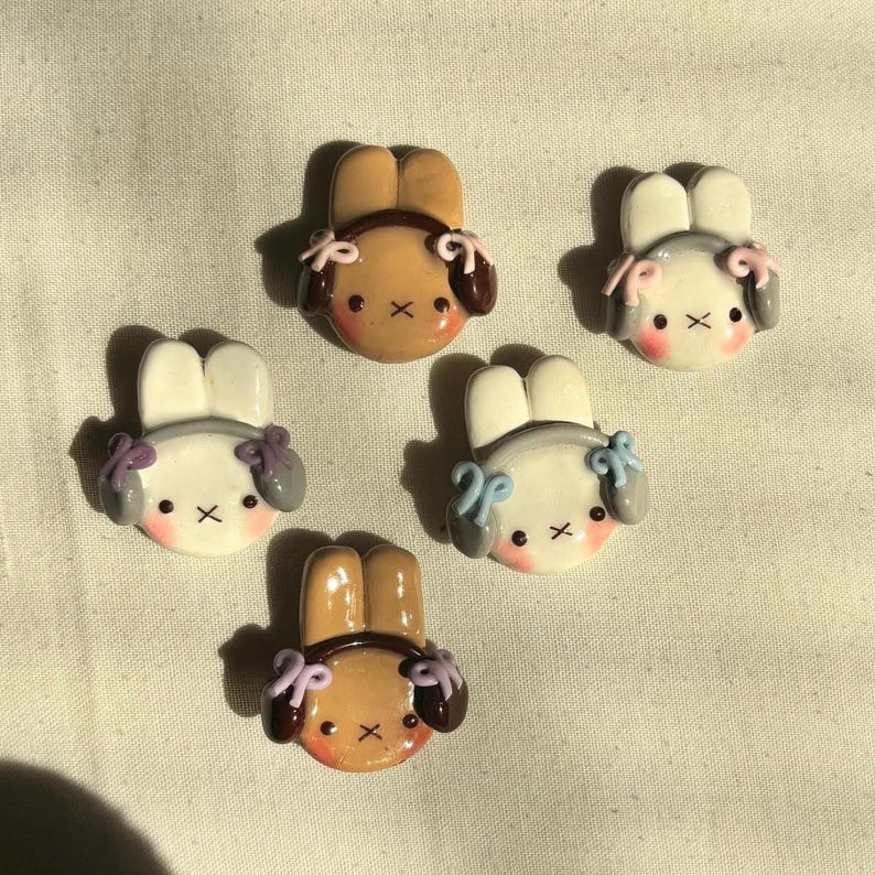 May include: Five handmade polymer clay bunny charms with pastel colored bows and gray or brown ears. The bunnies are wearing headphones.