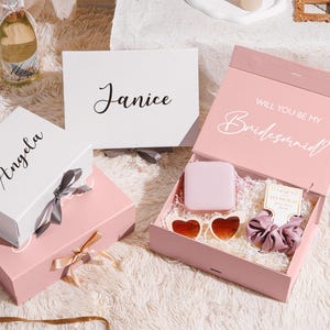 May include: A collection of gift boxes in white and pink, some with names like "Angela" and "Janice" printed on them. One box is open, revealing a gift set with heart-shaped sunglasses, a scrunchie, and a small square item. The open box reads "Will You Be My Bridesmaid?"
