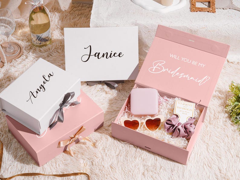 May include: A collection of gift boxes in white and pink, some with names like "Angela" and "Janice" printed on them. One box is open, revealing a gift set with heart-shaped sunglasses, a scrunchie, and a small square item. The open box reads "Will You Be My Bridesmaid?"