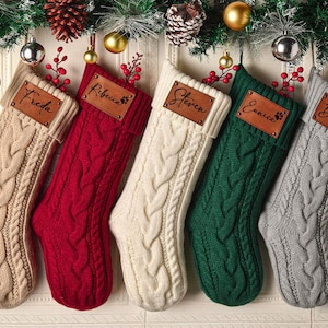 May include: Five cable-knit Christmas stockings in beige, red, cream, dark green, and gray. Each stocking features a rectangular brown leather patch with a name engraved in black script: Freda, Rebecca, Steven, Eunice, and Doris.
