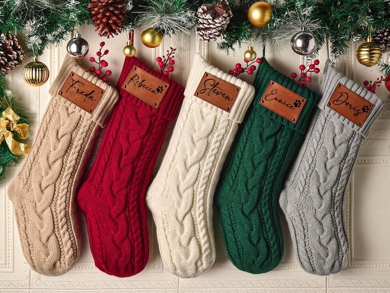 May include: Five cable-knit Christmas stockings in beige, red, cream, dark green, and gray. Each stocking features a rectangular brown leather patch with a name engraved in black script: Freda, Rebecca, Steven, Eunice, and Doris.