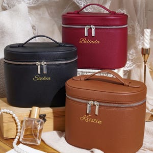 May include: Three oval-shaped cosmetic cases in black, burgundy, and brown with a textured finish. Each case features a top handle, a silver-toned double zipper, and a gold embroidered name: Sophia on the black, Belinda on the burgundy, and Kristin on the brown.