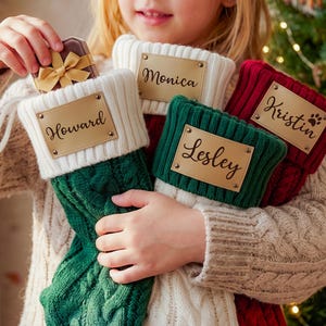 May include: Three cable-knit Christmas stockings in green, white, and red, each featuring a rectangular gold-toned metal nameplate. The plates are engraved with the names Howard, Monica, Kristin, and Lesley in a black script font.