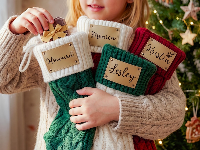 May include: Three cable-knit Christmas stockings in green, white, and red, each featuring a rectangular gold-toned metal nameplate. The plates are engraved with the names Howard, Monica, Kristin, and Lesley in a black script font.