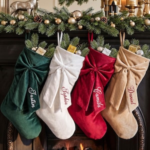 Könnte beinhalten: Vier Weihnachtssocken aus Samt in Dunkelgrün, Weiß, Rot und Beige, jeweils mit einer großen Schleife in der Mitte und den eingestickten Namen Justin, Sophia, Camila und Daniel. Die Socken sind mit Tannenzweigen und kleinen Geschenken gefüllt.