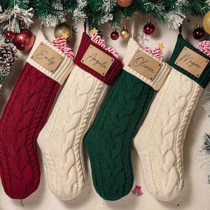 May include: Four knit Christmas stockings featuring a cable-knit pattern. Each stocking has a ribbed cuff with a rectangular leather-like tag attached. The tags are personalized with the names Emily, Angela, Oliver, and Megan in black script.