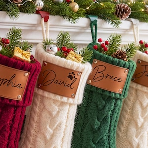 May include: Four cable-knit Christmas stockings in burgundy, cream, and forest green, each featuring a personalized tan leather patch with names Sophia, David, Bruce, and Janice, accented with pine sprigs, red berries, and small gift boxes.