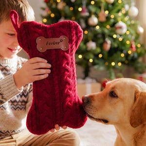 May include: A deep red, cable-knit Christmas stocking shaped like a dog bone. The stocking features a brown leather-like patch in the center with the name Amber and a small paw print icon engraved on it, along with a fabric loop for hanging.