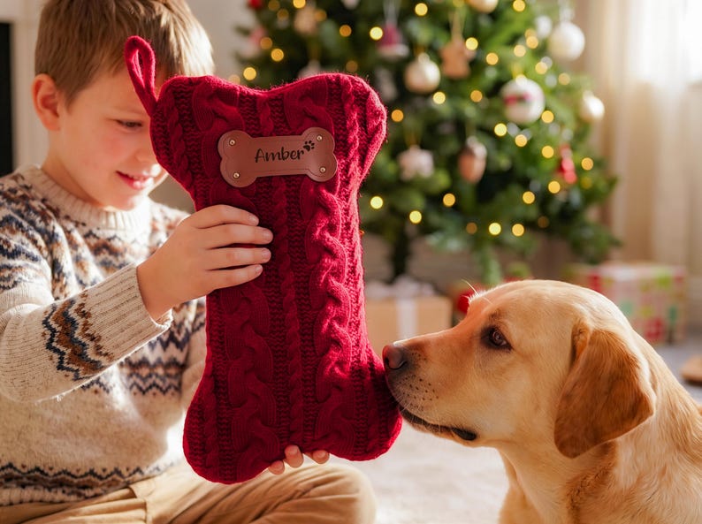 May include: A deep red, cable-knit Christmas stocking shaped like a dog bone. The stocking features a brown leather-like patch in the center with the name Amber and a small paw print icon engraved on it, along with a fabric loop for hanging.