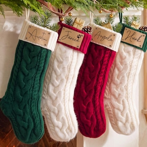 May include: Four cable-knit Christmas stockings in dark green, white, deep red, and white. Each stocking features a light-colored leather tag with the names Austin, Janice, Angela, and Hunk engraved in black script, topped with pine sprigs.
