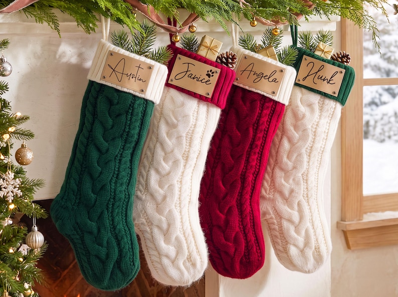 May include: Four cable-knit Christmas stockings in dark green, white, deep red, and white. Each stocking features a light-colored leather tag with the names Austin, Janice, Angela, and Hunk engraved in black script, topped with pine sprigs.