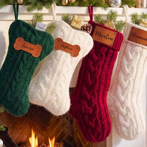 May include: Four cable-knit Christmas stockings in dark green, cream, burgundy, and white. The green and cream stockings feature bone-shaped leather tags with the names Louise and Sophia. The burgundy and white stockings have rectangular leather tags with the names Martin and Hannah.