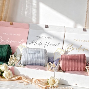 May include: Three gift boxes with the text "Will You Be My Bridesmaid?" and "Made of Honor?" Each box contains a corduroy clutch in green, gray, and pink. The boxes are decorated with ribbons, pearls, and cards.