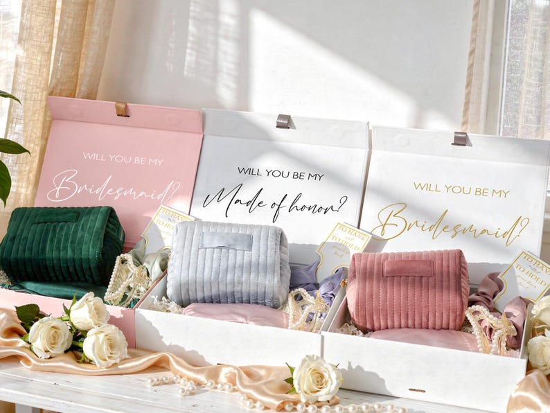 May include: Three gift boxes with the text "Will You Be My Bridesmaid?" and "Made of Honor?" Each box contains a corduroy clutch in green, gray, and pink. The boxes are decorated with ribbons, pearls, and cards.