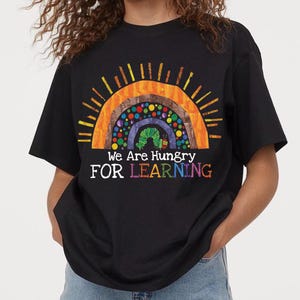 May include: Black t-shirt with a colorful graphic of a rainbow and a caterpillar. The text "We Are Hungry FOR LEARNING" is printed below the rainbow design. The shirt is a casual style.