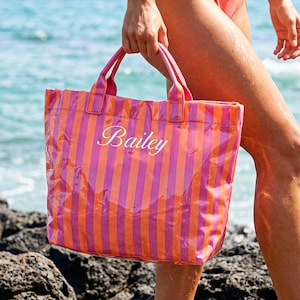 Könnte beinhalten: Eine rosa-orange gestreifte Tragetasche mit dem Namen "Bailey" in weißer Schrift. Die Tasche hat passende rosa Griffe. Die Tasche wird in der Nähe des Meeres gehalten. Die Tasche scheint aus einem transparenten, wasserdichten Material zu bestehen.