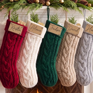 May include: Five cable-knit Christmas stockings in red, cream, dark green, beige, and light gray. Each stocking features a rectangular wooden name tag engraved with the names Kristin, Alexia, Jackson, David, and Hunk, attached to the cuff.
