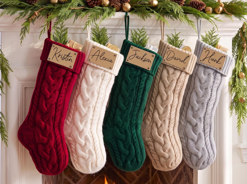 May include: Five cable-knit Christmas stockings in red, cream, dark green, beige, and light gray. Each stocking features a rectangular wooden name tag engraved with the names Kristin, Alexia, Jackson, David, and Hunk, attached to the cuff.