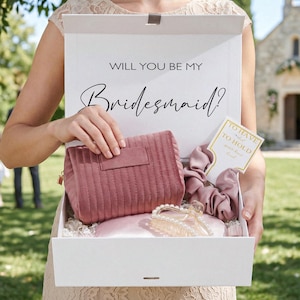 以下が含まれることがあります： 「WILL YOU BE MY Bridesmaid?」の文字が入った白い箱には、ピンクの化粧ポーチ、シュシュ、ヘアクリップ、カードが入っています。明るい色のドレスを着た人が箱を持っており、背景には教会があります。
