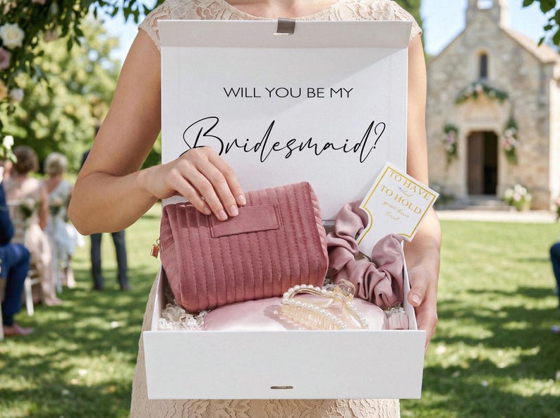 May include: A white box with the text "WILL YOU BE MY Bridesmaid?" contains a pink cosmetic bag, scrunchies, a hair clip, and a card. The box is held by a person in a light-colored dress, with a church in the background.