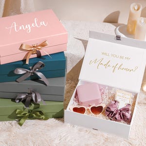 May include: A stack of gift boxes in various colors, including pink, teal, gray, and green, with satin ribbon bows. The top box is pink with the name "Angela" in script. An open white box reveals gifts, including heart-shaped sunglasses and a scrunchie. Text reads "Will you be my Made of honor?"