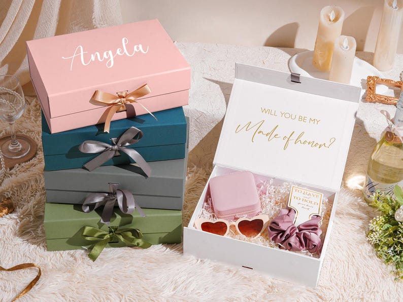 May include: A stack of gift boxes in various colors, including pink, teal, gray, and green, with satin ribbon bows. The top box is pink with the name "Angela" in script. An open white box reveals gifts, including heart-shaped sunglasses and a scrunchie. Text reads "Will you be my Made of honor?"