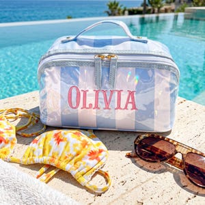 Puede incluir: Un neceser rectangular con un patrón de rayas azul claro y blanco y un acabado iridiscente. El nombre OLIVIA está bordado en rosa en la parte frontal. El bolso cuenta con un asa superior y un cierre de doble cremallera. Un top de bikini floral amarillo y unas gafas de sol marrones están colocados cerca.