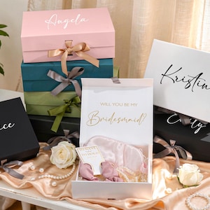 May include: A collection of gift boxes in various colors, including pink, teal, black, and white. Each box is adorned with a ribbon and a personalized name in elegant script. One box is open, revealing a satin sleep mask and other gifts. The text "Will you be my Bridesmaid?" is visible.