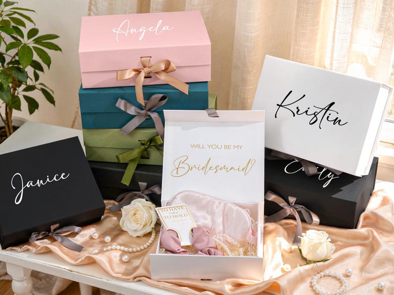 May include: A collection of gift boxes in various colors, including pink, teal, black, and white. Each box is adorned with a ribbon and a personalized name in elegant script. One box is open, revealing a satin sleep mask and other gifts. The text "Will you be my Bridesmaid?" is visible.