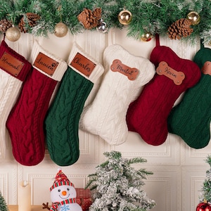 May include: Six Christmas stockings in white, red, and green, with a cable knit design. Each stocking has a brown leather tag with a name on it. Two stockings are shaped like dog bones. The stockings are suspended from a festive garland.