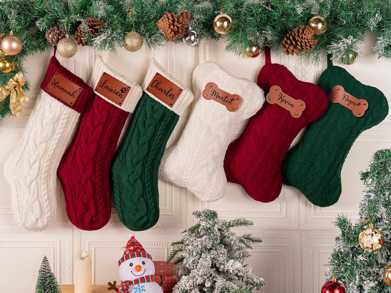 May include: A collection of six Christmas stockings hanging in a row. Three are traditional boot-shaped stockings in cream, red, and green with rectangular leather name tags. Three are bone-shaped stockings in cream, red, and green with bone-shaped leather tags.
