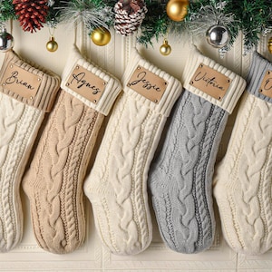 May include: Five cable-knit Christmas stockings in cream, beige, and grey, each featuring a ribbed cuff and a rectangular leather-like patch. The patches are personalized with the names Brian, Agnes, Jessie, Victoria, and Odelia in black script.