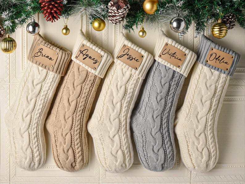 May include: Five cable-knit Christmas stockings in cream, beige, and grey, each featuring a ribbed cuff and a rectangular leather-like patch. The patches are personalized with the names Brian, Agnes, Jessie, Victoria, and Odelia in black script.