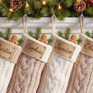 May include: Four cream-colored cable knit Christmas stockings hanging in a row. Each stocking features a rectangular gold-toned metal nameplate engraved with the names Martin, Sophia, Janice, and Oliver, topped with a small gingerbread man.