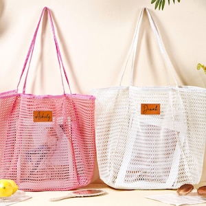 以下が含まれることがあります： ピンクと白のメッシュトートバッグ2つ。長いハンドルが付いています。ピンクのバッグには「Ashely」という名前が茶色の革パッチに、白いバッグには「Dinah」という名前が付いています。ビーチやショッピングに最適です。