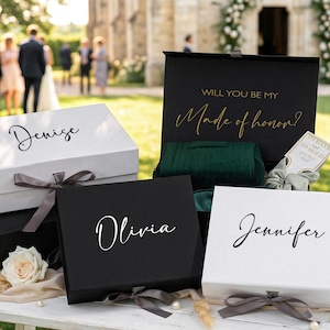 May include: Several gift boxes, including white and black, are displayed outdoors. The boxes are tied with gray ribbons and have names like "Denise," "Olivia," and "Jennifer" written on them. One box says "Will you be my Made of honor?"