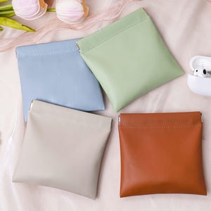 Può includere: Quattro piccole pochette quadrate in colori pastello: azzurro, verde, beige e marrone. È visibile anche una custodia bianca con auricolari e un singolo auricolare. Le pochette sono probabilmente per piccoli oggetti.
